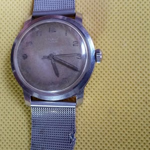 May include: A vintage silver-toned wristwatch with a mesh band. The watch face is a light color with black numerals and hands. The watch is set against a yellow background.