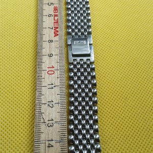 Puede incluir: Una correa de reloj plateada con un diseño de malla, junto a una regla de madera. La regla muestra medidas en centímetros, con la correa de reloj midiendo aproximadamente 15 cm de largo.