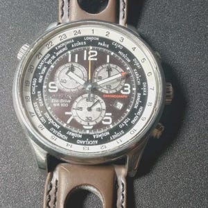 May include: A Citizen Eco-Drive WR 100 chronograph watch with a brown dial and a brown leather strap. The watch has multiple subdials, a world time bezel, and a stainless steel case.