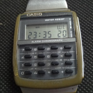 May include: A vintage Casio digital watch with a calculator keypad. The watch face displays the time and day. The watch has a silver metal band and a green and silver watch face. The watch is labeled "CASIO" and "WATER RESIST".