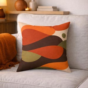 May include: A decorative throw pillow with an abstract design. The pillow features a beige background with layered shapes in shades of orange, brown, and green. The pillow is placed on a light-colored sofa.