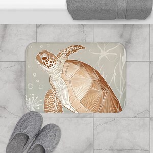 Puede incluir: Una alfombra de baño rectangular con una ilustración detallada de una tortuga marina en tonos marrones y beige. La tortuga está colocada sobre un fondo gris claro con detalles de plantas acuáticas y burbujas. Un par de zapatillas de punto grises en primer plano.