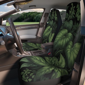 以下が含まれることがあります： 黒い背景に、緑豊かな熱帯の葉のデザインが施された車のシートカバー。運転席と助手席のシートにカバーが装着されており、ハンドルとダッシュボードが見えます。デザインには、大きく詳細な葉が含まれています。