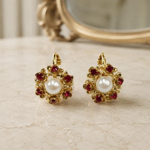 May include: A pair of gold-tone earrings featuring a central white faux pearl surrounded by a circular arrangement of small, vibrant red faceted crystals. The earrings have a secure lever-back closure mechanism and a polished metallic finish.