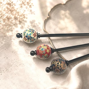 Può includere: Tre bastoncini per capelli con lunghi steli neri e cime sferiche. Le sfere superiori presentano intricati motivi floreali in bianco, rosso e blu scuro, ciascuno accentato con dettagli dorati e rifinito con una piccola perla nera all'estremità.