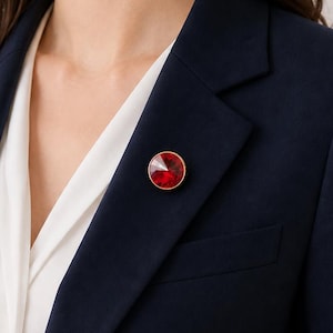 Pode incluir: Uma broche redonda de pedra preciosa vermelha com uma borda dourada é presa a um blazer azul marinho. A broche é o ponto focal, adicionando um toque de cor contra o tecido escuro. Uma blusa branca é visível por baixo.