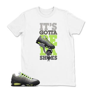 May include: White t-shirt with the text "IT'S GOTTA BE DA SHOES" in gray and green. The graphic features a cartoon character holding a gray and neon green sneaker. A matching sneaker is shown at the bottom of the image.
