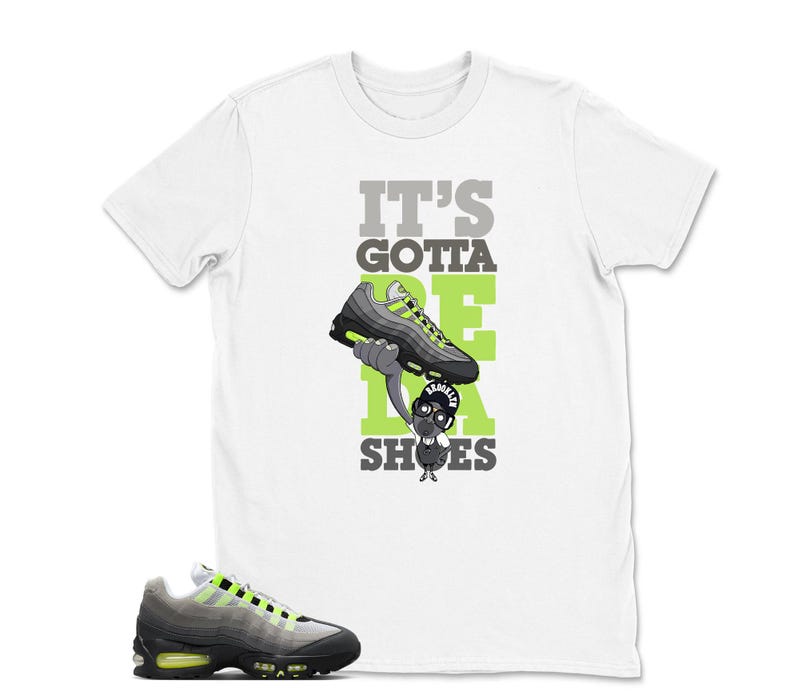 May include: White t-shirt with the text "IT'S GOTTA BE DA SHOES" in gray and green. The graphic features a cartoon character holding a gray and neon green sneaker. A matching sneaker is shown at the bottom of the image.