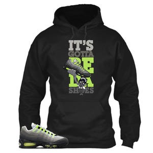May include: Black hooded sweatshirt with the text "IT'S GOTTA BE DA SHOES" in gray and green. The graphic features a cartoon character holding a sneaker. A gray and neon green sneaker is in the lower left corner.