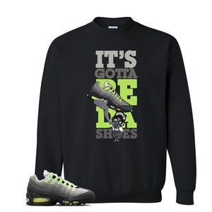 May include: Black sweatshirt with the text "IT'S GOTTA BE DA SHOES" in gray and neon green. The graphic features a cartoon character holding a neon green and gray sneaker. A matching sneaker is in the foreground.