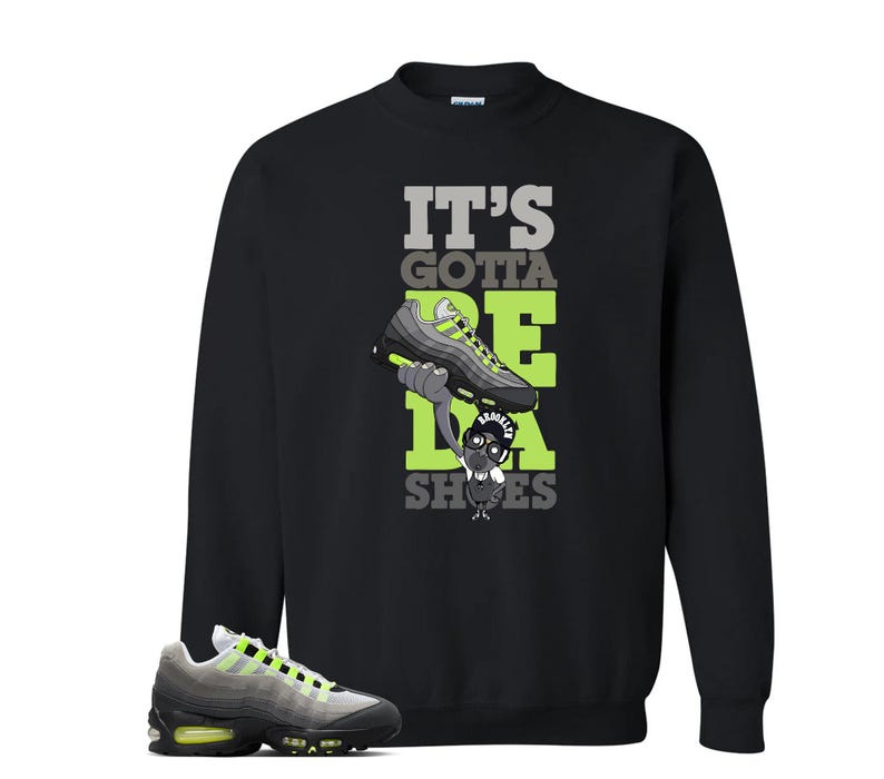 May include: Black sweatshirt with the text "IT'S GOTTA BE DA SHOES" in gray and neon green. The graphic features a cartoon character holding a neon green and gray sneaker. A matching sneaker is in the foreground.