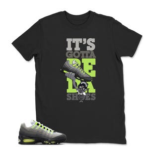 May include: Black t-shirt with the text "IT'S GOTTA BE DA SHOES" in gray and neon green. The graphic features a cartoon character holding a sneaker. A matching gray and neon green sneaker is in the foreground.