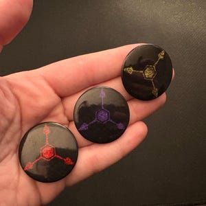 May include: Three black round buttons with geometric designs in red, purple, and gold. The buttons are held in a hand, showcasing the different color variations of the design. The buttons are likely for clothing or accessories.