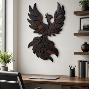 May include: A wall-mounted wooden sculpture of a phoenix with wings spread upward. The piece is constructed from individual segments of wood in varying shades of dark charcoal, black, and deep reddish-brown, creating a layered, textured effect.