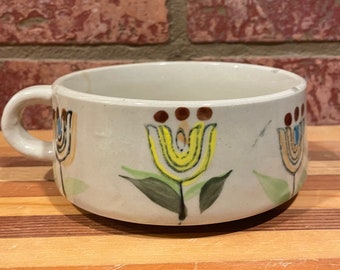 May include: A white ceramic soup bowl with a handle, featuring a stylized floral design in yellow, brown, and green. The bowl has a rounded shape and a slightly flared rim.