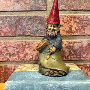 May include: A whimsical garden gnome figurine with a red pointed hat, gray sweater, and green leaf-shaped skirt. The gnome holds a paintbrush and a palette. The figurine is resting on a vintage blue book.