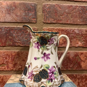 May include: A white porcelain creamer with a handle and spout, adorned with purple flowers, teal accents, and gold trim. The creamer has a decorative floral and pinecone design. The creamer is sitting on a blue book.