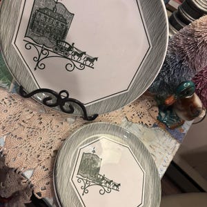 May include: Two white octagonal plates with a black and gray rim. Each plate features a black illustration of a building and horse-drawn carriage. The plates are displayed on a black metal stand.