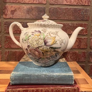 May include: A white ceramic teapot with a floral design in shades of purple, yellow, and green. The teapot is placed on top of two antique books with blue and red covers, against a brick wall background.