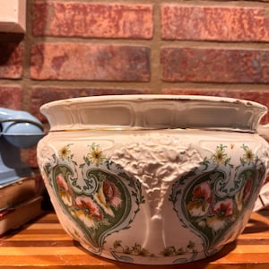 May include: A white ceramic chamber pot with a decorative floral design in green, pink, and yellow. The pot has a handle and a gold trim around the rim. The background includes a brick wall and a vintage blue telephone.