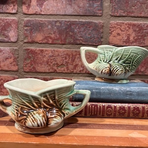 May include: Two vintage ceramic planters with a green and brown leaf design. One planter has handles, and the other has a spout. They are displayed on top of two antique books with a brick background.
