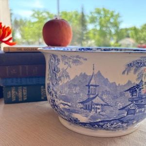 May include: A white ceramic chamber pot featuring a blue transferware pattern depicting a landscape with buildings, trees, and mountains. The vessel has a single handle on the right side and a wide, circular rim, showcasing a detailed scene.