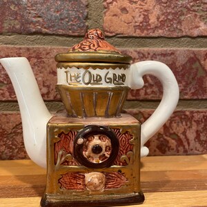 May include: A white ceramic teapot shaped like an old-fashioned coffee grinder. The teapot has a brown and gold design with the words "The Old Grind" on the front.