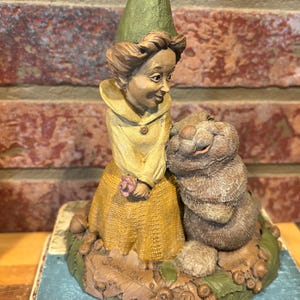 May include: A whimsical figurine of a woman in a yellow dress and green hat, with a friendly-looking brown rabbit. The base is decorated with leaves and acorns. The figurine is set against a brick wall.