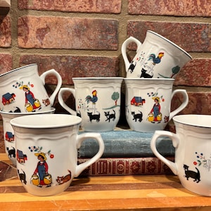 May include: A collection of six white ceramic mugs featuring a repeating pattern of a person carrying milk pails, black cats, a rooster on a fence, and a red barn. Each mug has a scalloped rim and a dark blue line along the top edge.