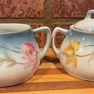 May include: A vintage porcelain creamer and sugar set with a blue and white ombre glaze. The set features a floral design with yellow and pink flowers.