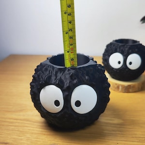 Op de afbeelding: Zwarte, bolvormige plantenbak met grote witte ogen, die op een stripfiguur lijkt. De plantenbak is ongeveer 9 cm hoog. Een gele meetlint is in de plantenbak geplaatst. Een andere vergelijkbare plantenbak staat op de achtergrond.