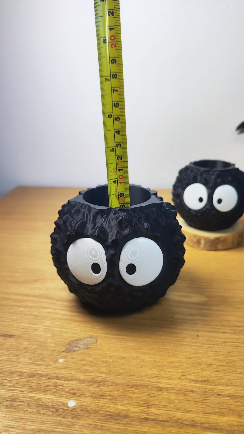 Op de afbeelding: Zwarte, bolvormige plantenbak met grote witte ogen, die op een stripfiguur lijkt. De plantenbak is ongeveer 9 cm hoog. Een gele meetlint is in de plantenbak geplaatst. Een andere vergelijkbare plantenbak staat op de achtergrond.