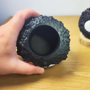 Op de afbeelding: Een zwarte, getextureerde, ronde container met een grote opening. De container heeft een ruw, ongelijkmatig oppervlak. Het object staat op een houten oppervlak.