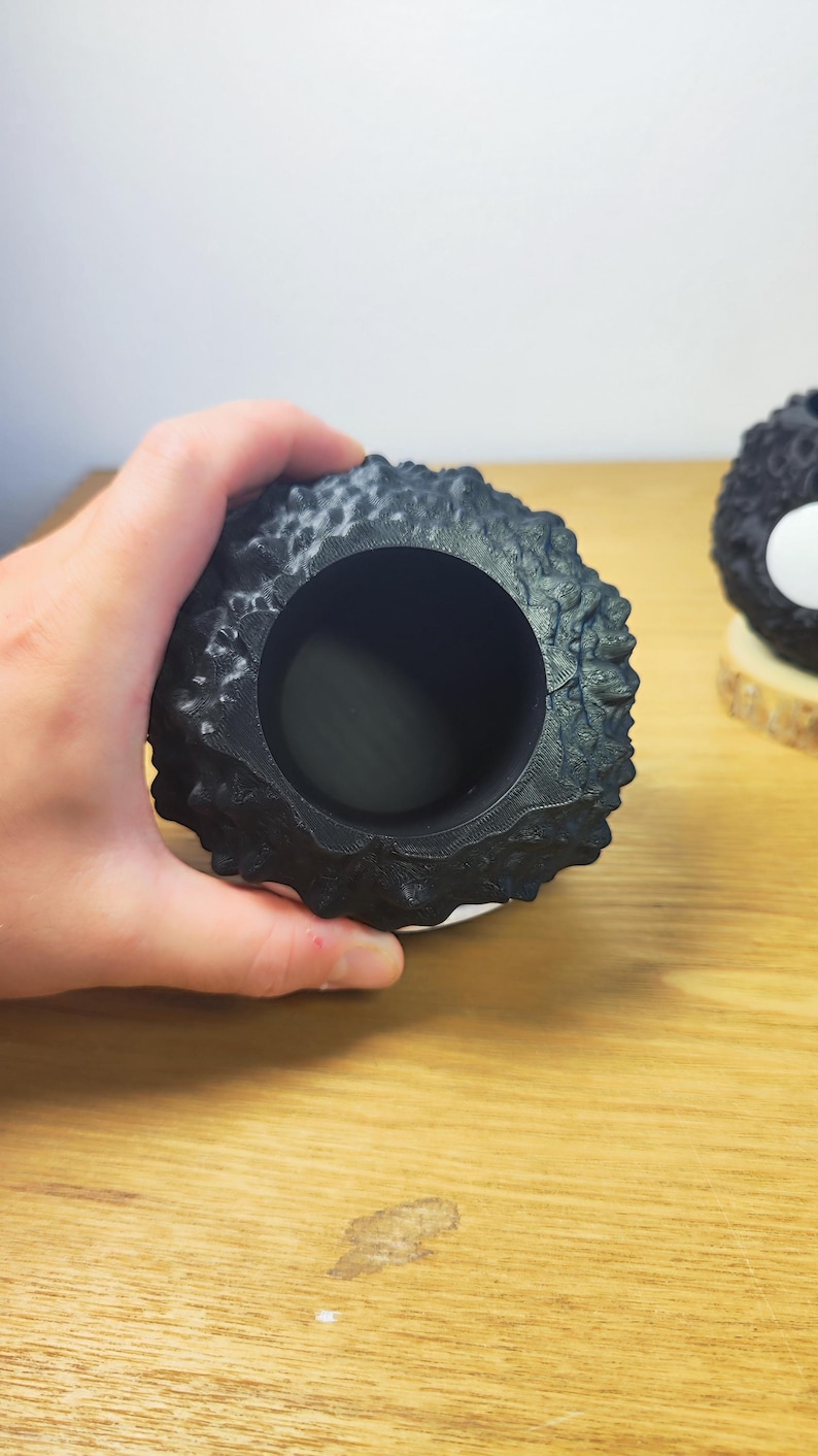 Op de afbeelding: Een zwarte, getextureerde, ronde container met een grote opening. De container heeft een ruw, ongelijkmatig oppervlak. Het object staat op een houten oppervlak.