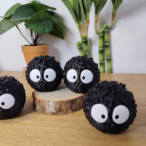 May include: Four black, round, fuzzy figures with large white eyes. They are arranged on a wooden surface, with a small wooden slice beneath two of them. A potted plant and bamboo stalks are in the background.