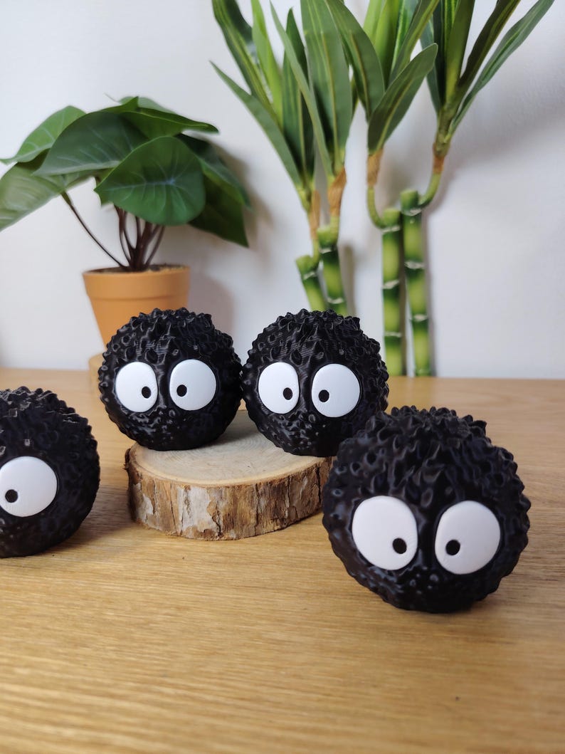 May include: Four black, round, fuzzy figures with large white eyes. They are arranged on a wooden surface, with a small wooden slice beneath two of them. A potted plant and bamboo stalks are in the background.