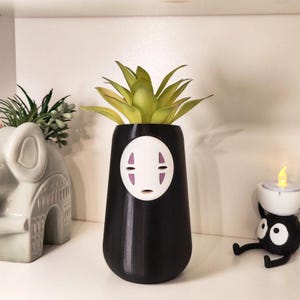 Puede incluir: Un jarrón negro con un diseño de cara blanca, que contiene una planta suculenta verde y amarilla. El jarrón tiene forma redondeada y está colocado sobre una estantería blanca. También se ven objetos decorativos, como una pequeña maceta con forma de elefante y un portavelas de duende de hollín.