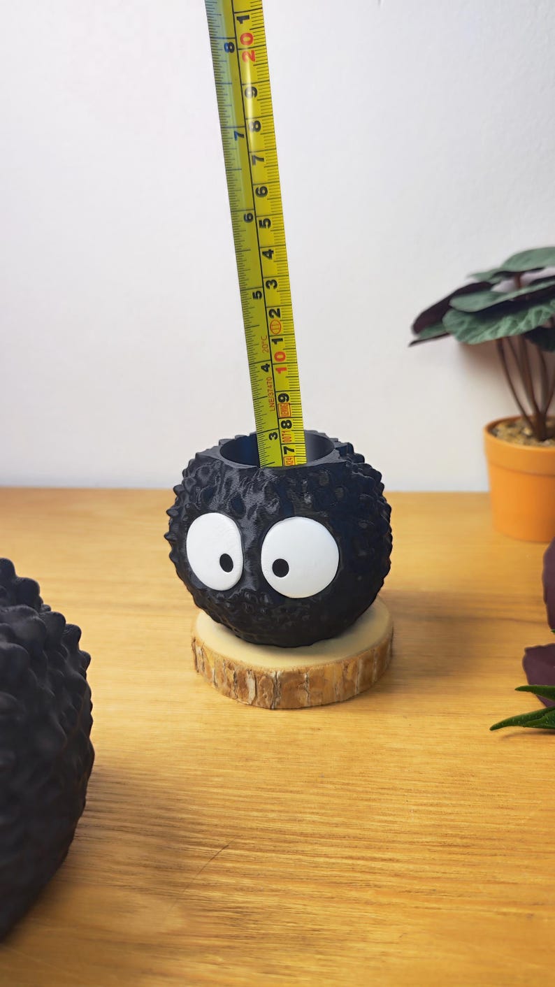Op de afbeelding: Een zwarte, getextureerde, bolvormige plantenbak met grote witte ogen, die lijkt op een roetelf. Hij staat op een kleine houten schijf. De plantenbak heeft een diameter van ongeveer 7.6 cm. Op een houten oppervlak.