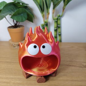 Può includere: Un contenitore a forma di fuoco di cartone animato, stampato in 3D in rosso e arancione, con una base marrone a forma di tronco. Presenta grandi occhi bianchi e un'ampia apertura. Il design è stravagante e decorativo.