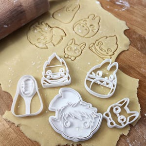 Puede incluir: Una colección de cortadores de galletas de plástico blanco con varias formas de personajes de dibujos animados, incluido un diseño de Totoro. Los cortadores están colocados sobre una lámina de masa, con un rodillo de madera en el fondo. La masa está ligeramente espolvoreada con harina.