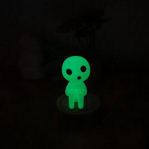 May include: A green glow-in-the-dark figurine of a small, round, humanoid character with two eyes and a mouth. The figurine is standing on a small, round base.