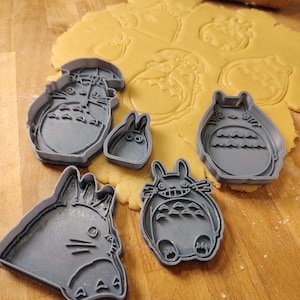 Pode incluir: Cortadores de biscoitos de plástico cinza em várias formas do personagem animado Totoro e um coelho pequeno. Os cortadores estão em uma superfície de madeira com massa aberta e um rolo no fundo. Tema de panificação e cozinha.