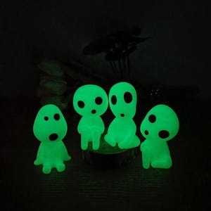 May include: Four glowing green figurines with round eyes and mouths, resembling characters from the animated film 'My Neighbor Totoro'. The figurines are sitting on a wooden surface.