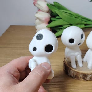 K&ouml;nnte beinhalten: Drei wei&szlig;e Figuren mit schwarzen Augen und Flecken, die an Figuren aus dem Film "Mein Nachbar Totoro" erinnern. Die Figuren stehen auf einem Holzsockel, eine Figur wird in der Hand gehalten.