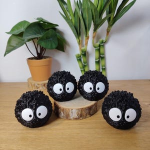 May include: Four black, round, fuzzy decorative figures with large white eyes. They are arranged on a wooden surface, with a small potted plant and bamboo stalks in the background. The figures resemble soot sprites.