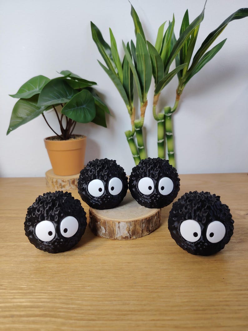 May include: Four black, round, fuzzy decorative figures with large white eyes. They are arranged on a wooden surface, with a small potted plant and bamboo stalks in the background. The figures resemble soot sprites.