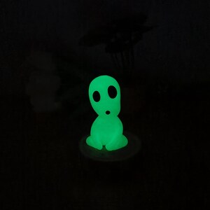 May include: A green glow-in-the-dark figurine of a character from the movie 'My Neighbor Totoro'. The figurine is sitting on a small wooden base.