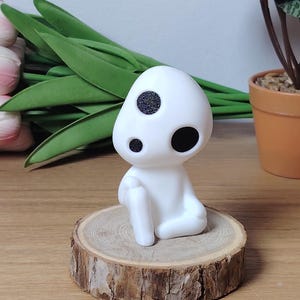 K&ouml;nnte beinhalten: Wei&szlig;e Figur eines Charakters mit zwei schwarzen Augen, sitzend auf einer Holzscheibe. Die Figur ist ein beliebter Charakter aus dem Animationsfilm 'Howls bewegliches Schloss'.