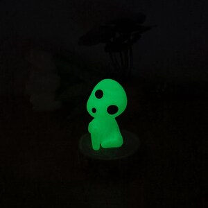 May include: A green glow-in-the-dark figurine of a mushroom-like creature with black dots. The figurine is sitting on a small wooden base.