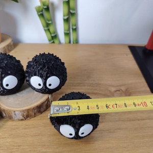 May include: Three black, round, spiky figurines with large white eyes. A measuring tape shows one figurine is approximately 5 cm in diameter. The figurines are on a wooden surface.